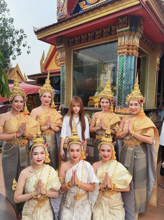 Thai dancers in gold and cream costumes with tall golden headdresses posing with the event host in front of a decorated Thai shrine