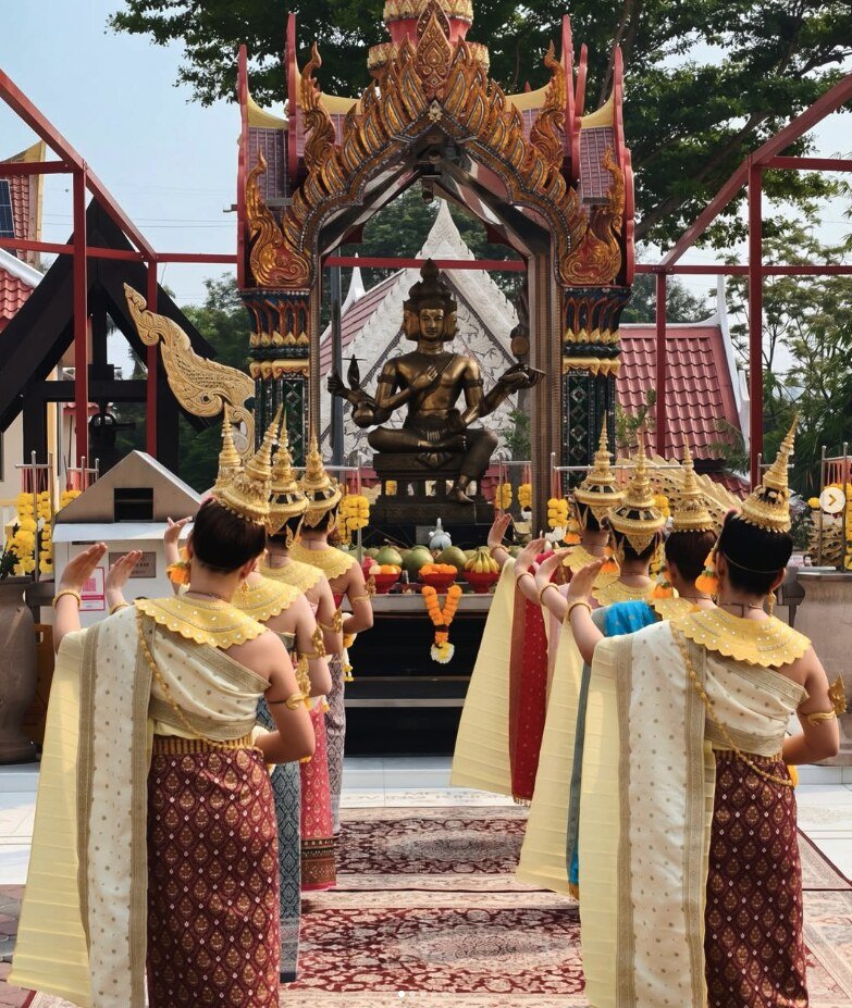 A row of Thai dancers in gold headdresses and silk costumes performing an offering dance in front of a Four-Faced Buddha shrine